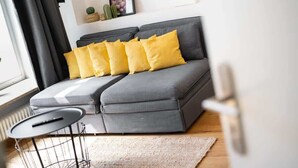 Ein modernes Wohnzimmer mit einer grauen Couch, gelben Kissen, einem kleinen runden Tisch und Blick ins Freie.