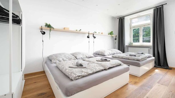 Ein modernes Schlafzimmer mit zwei Betten, Holzboden, Fenster mit Vorhängen und einer Pflanzenregal.