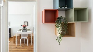 Ein Zimmer mit einem weißen Tisch und Stühlen, einer Pflanze in einer Holzschachtel und drei farbenfrohen Regalen an der Wand.