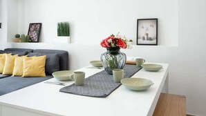 Ein modernes Wohnzimmer mit einem weißen Sofa, gelben Kissen, einem Couchtisch mit Blumen und einem Essbereich mit Tisch, Schüsseln und Tassen.