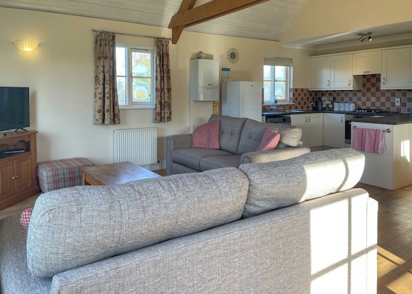 A living room with a sofa, a television, a small table, and a kitchen area with cabinets and a stove. - 2 bedroom accommodation in Palgrave, Diss (Palgrave, Diss)