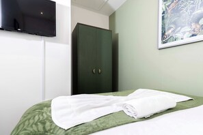 Ein Hotelzimmer mit einem Bett, einer grünen Bettdecke, einem weißen Handtuch, einem dunkelgrünen Kleiderschrank und einem flachbildfernseher an der Wand.