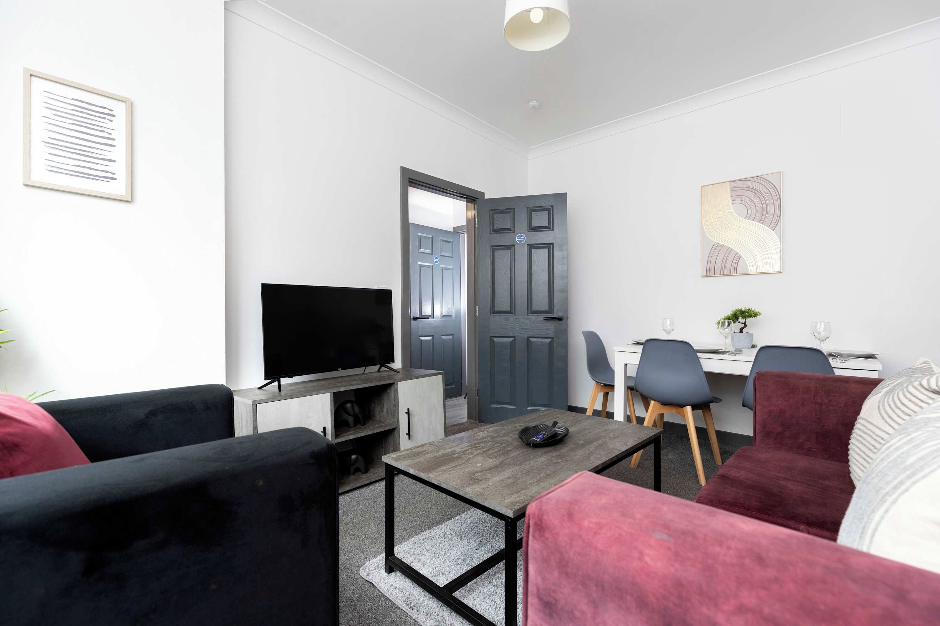A modern living room with a flat-screen TV, a dining area with a table and chairs, and a grey door.
