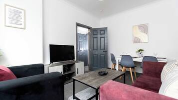 A modern living room with a flat-screen TV, a dining area with a table and chairs, and a grey door.