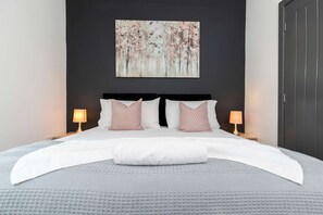 Ein ordentlich bezogenes Bett mit weißer Daunendecke, zwei Kissen und einer passenden Tagesdecke. Auf jeder Seite des Bettes stehen zwei Nachttischlampen.