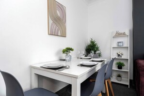 A dining area with a white table set for four, surrounded by chairs, with a bookshelf and a decorative piece on the wall.