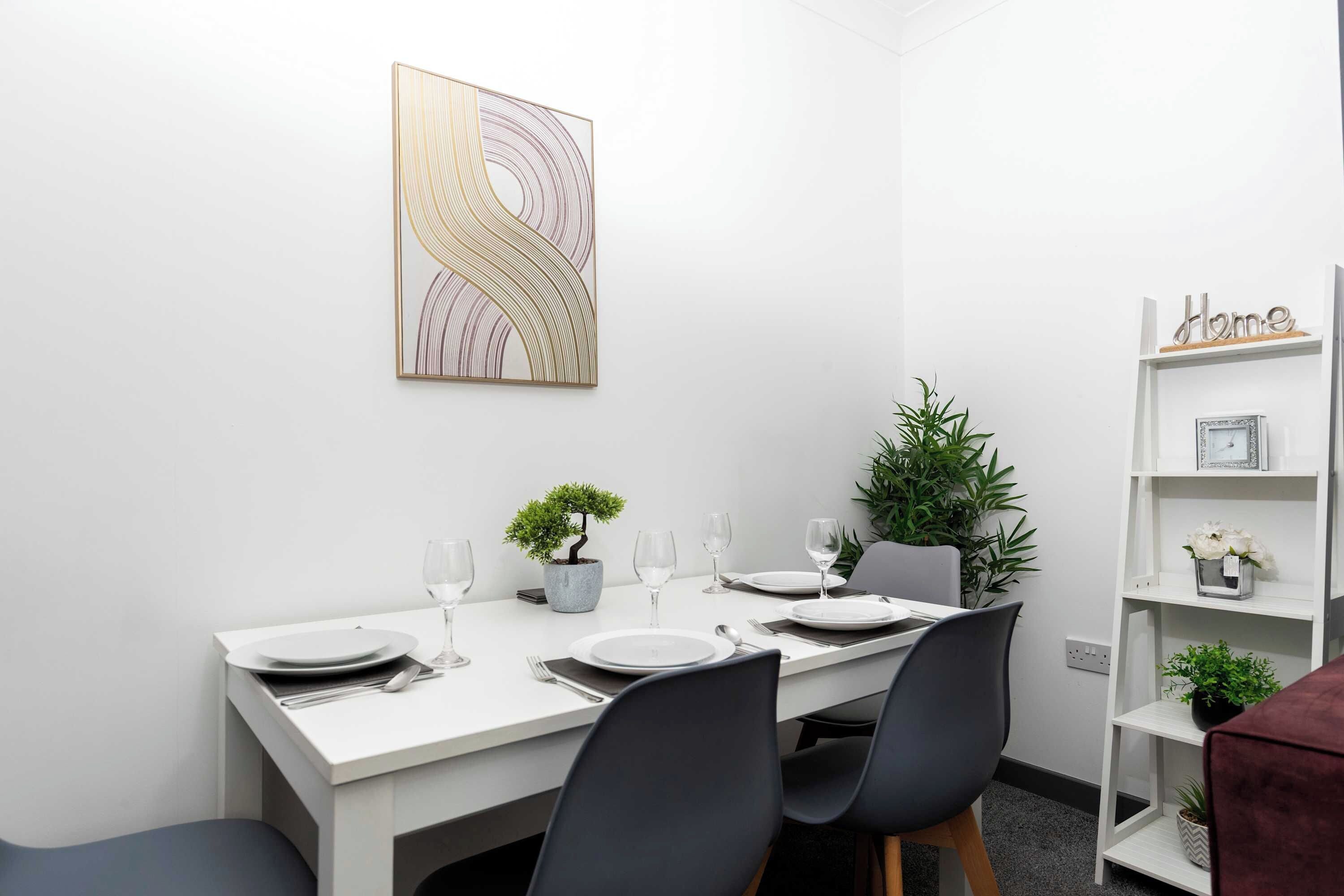 A dining area with a white table set for four, a potted plant, and a decorative wall piece.