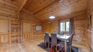 L’intérieur d’un chalet en bois comprend une table à manger, des chaises, un luminaire suspendu et une fenêtre avec des rideaux.