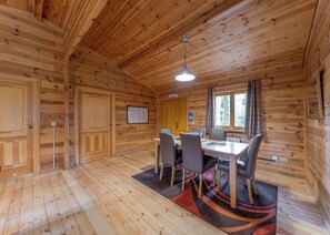A wooden cabin interior with a dining table, chairs, a hanging light fixture, and a window with curtains.