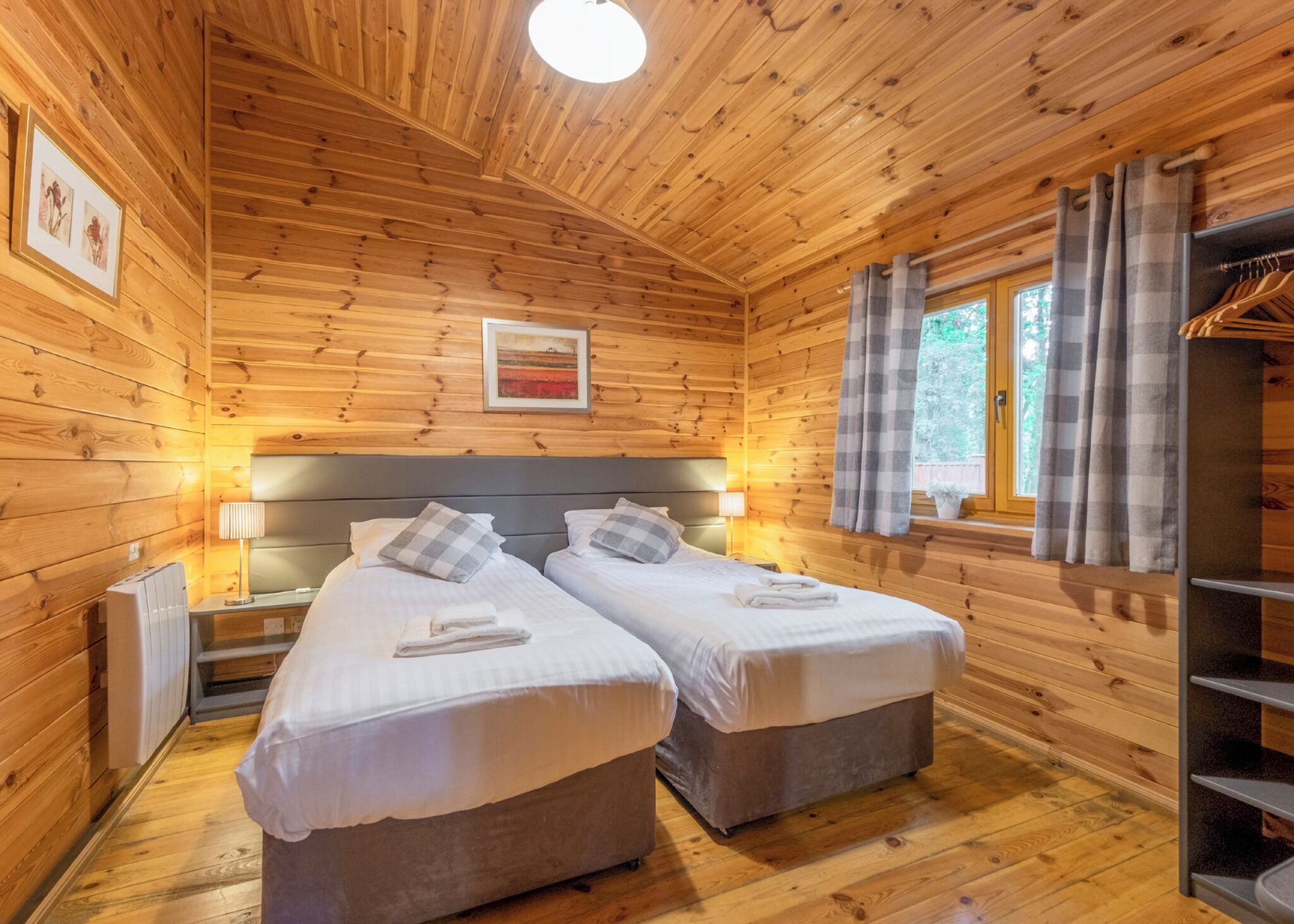 A wooden cabin room with two beds, a window with curtains, a radiator, and a framed picture on the wall.
