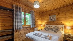 A wooden cabin bedroom with a bed, two bedside lamps, a window with curtains, and a painting on the wall. - 3 bedroom accommodation in Ellesmere (Ellesmere)