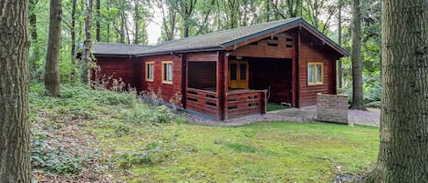 En enkel stuga med en veranda, omgiven av höga träd och en gräsyta.