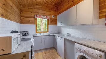 A compact kitchen with wooden ceiling, white cabinets, a window with a view of trees, a washing machine, and a microwave.