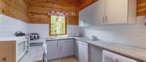 A compact kitchen with wooden ceiling, white cabinets, a window with a view of trees, a washing machine, and a microwave.