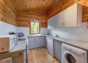 Une cuisine compacte avec un plafond en bois, des placards blancs, une fenêtre donnant sur les arbres, un lave-linge et un four à micro-ondes.