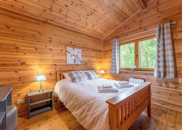 A wooden cabin bedroom with a bed, bedside lamps, a window with curtains, and a wooden headboard. - 3 bedroom accommodation in Ellesmere (Ellesmere)