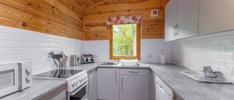 A compact kitchen with wooden ceiling, white tiled walls, a window with a floral curtain, and basic appliances.