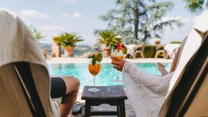 Deux personnes sont assises au bord d’une piscine, des boissons décorées de fruits sont posées devant elles, et elles sont entourées de palmiers et de chaises longues.