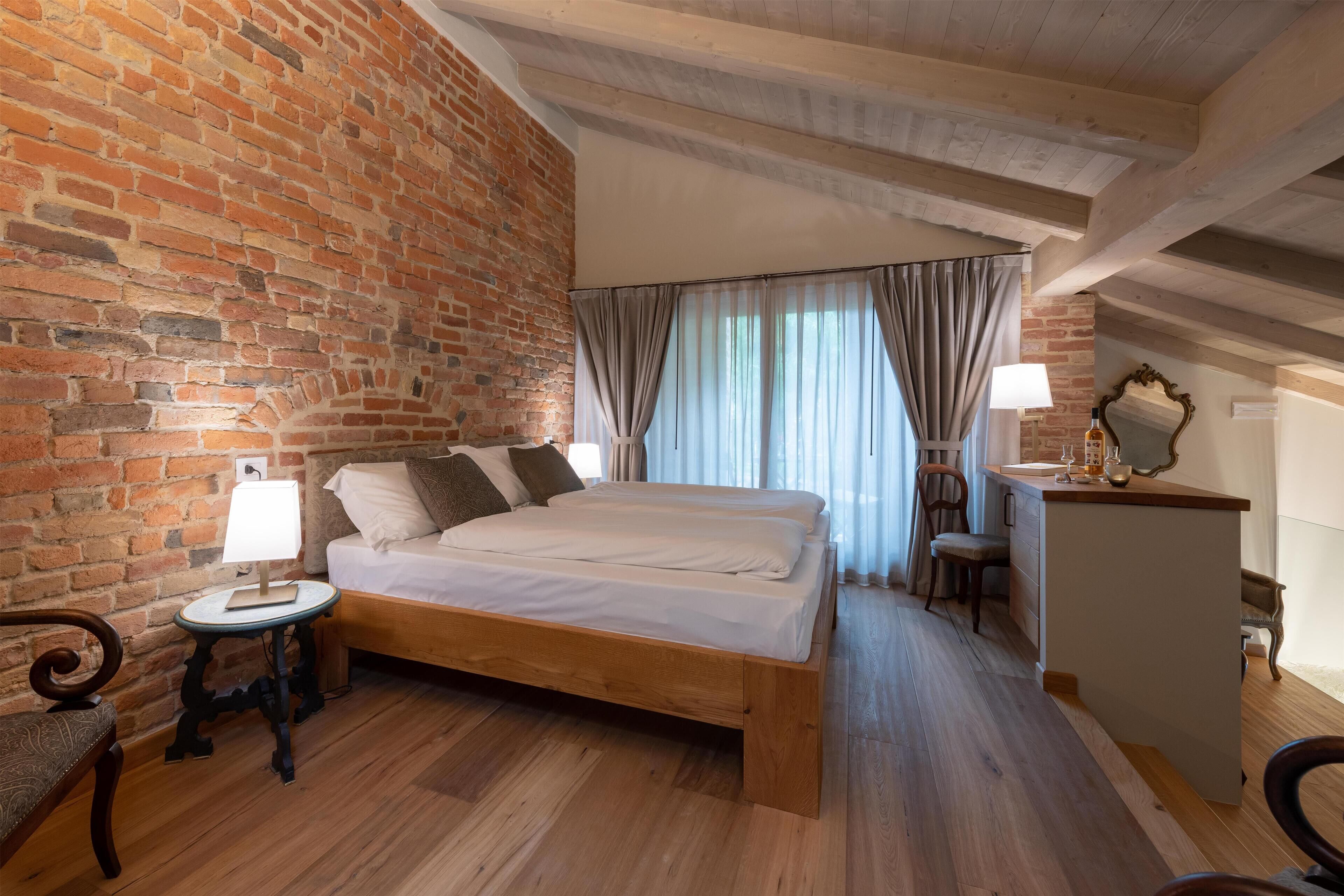 A bedroom with a wooden bed, a brick wall, a small bedside table, a lamp, a chair, and a desk with a mirror.