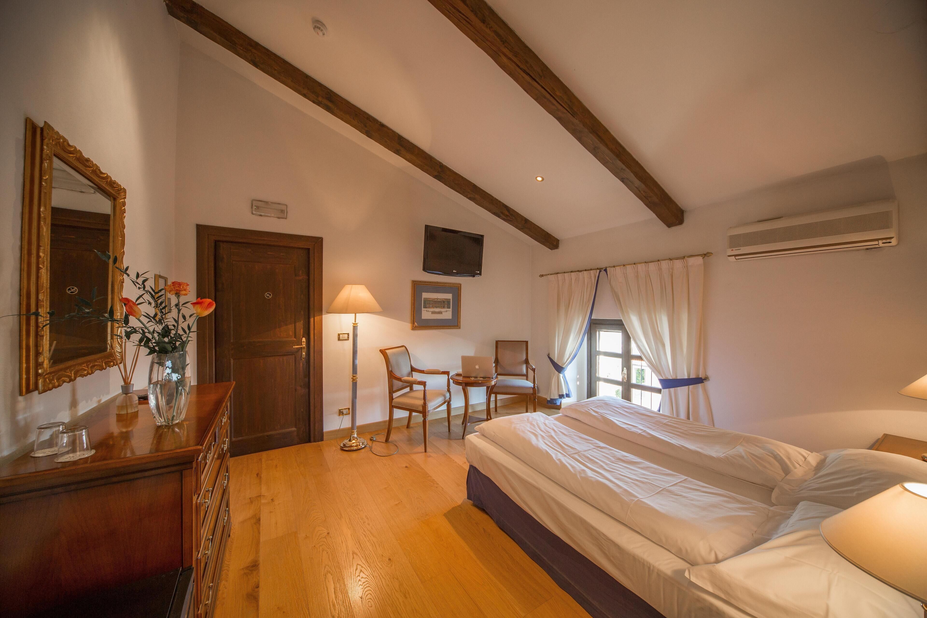 A hotel room with a wooden floor, a bed, a desk with a chair, a mirror, a vase with flowers, and an air conditioning unit.