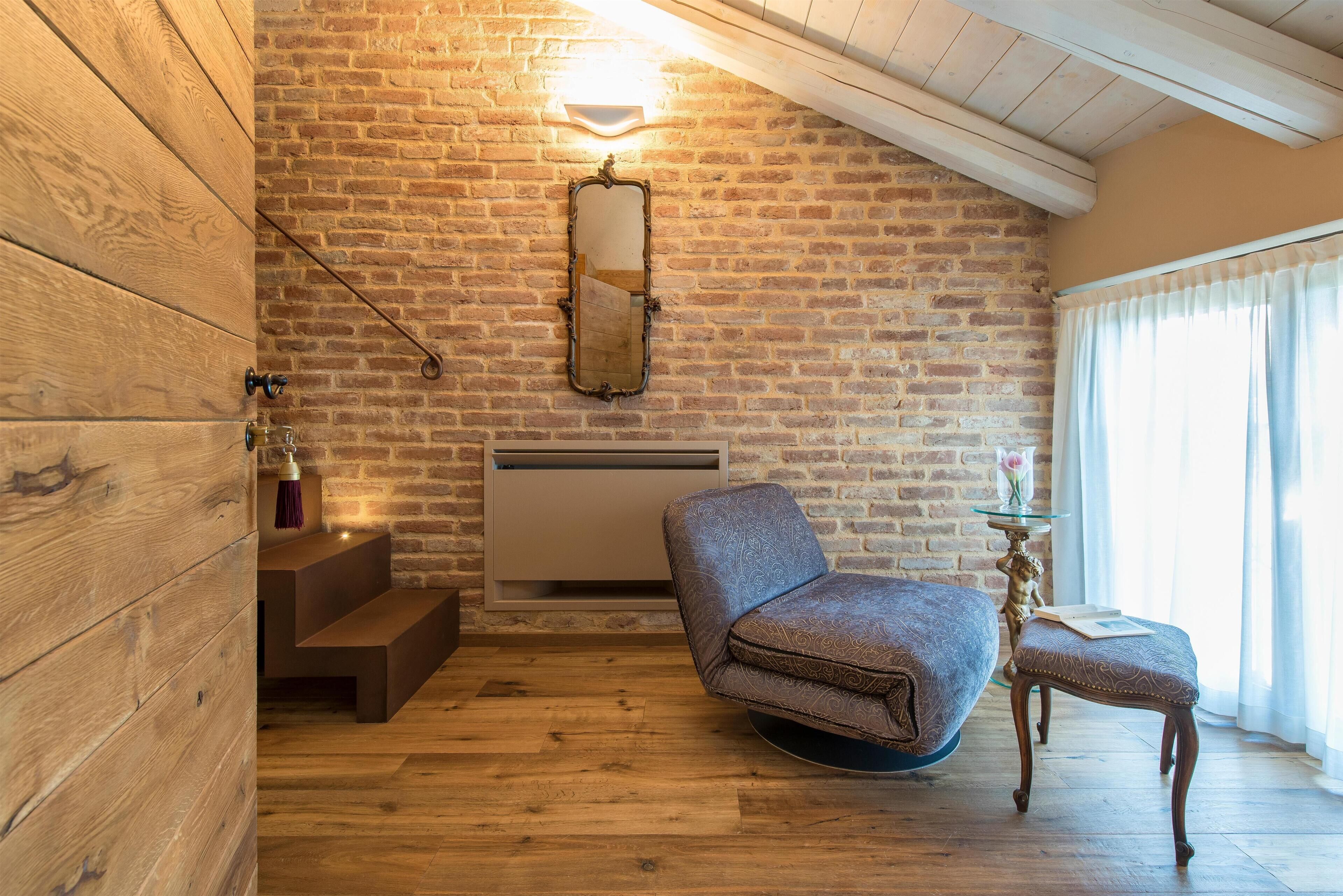 A room with a brick wall, wooden flooring, a chair, a small table, and a mirror.