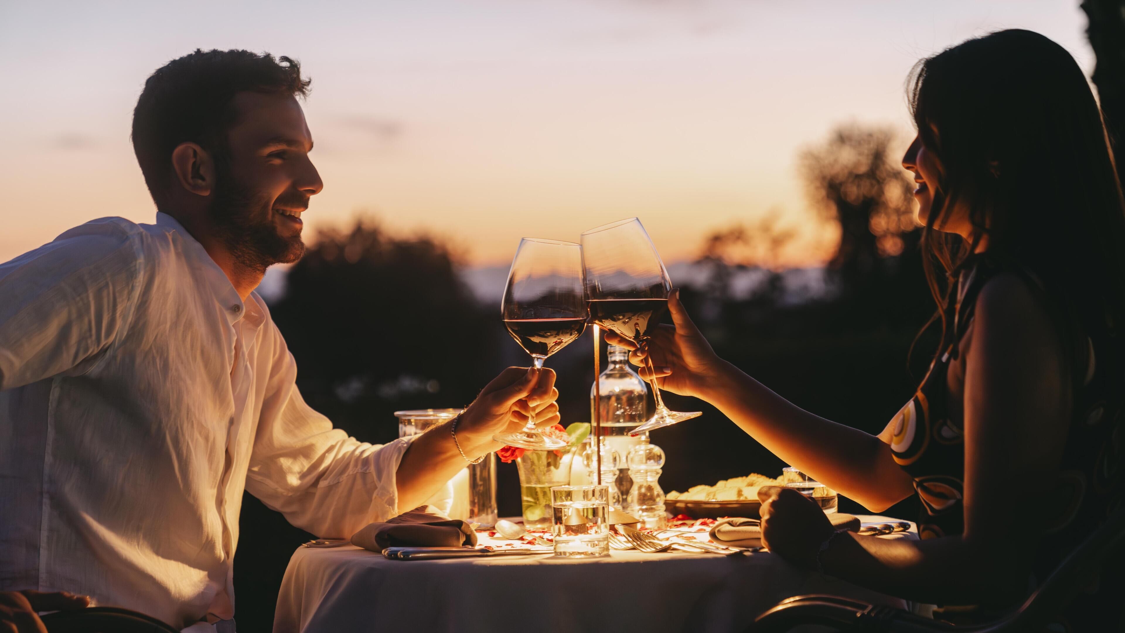 Um casal brindando com taças de vinho em uma mesa ao ar livre durante o pôr do sol.