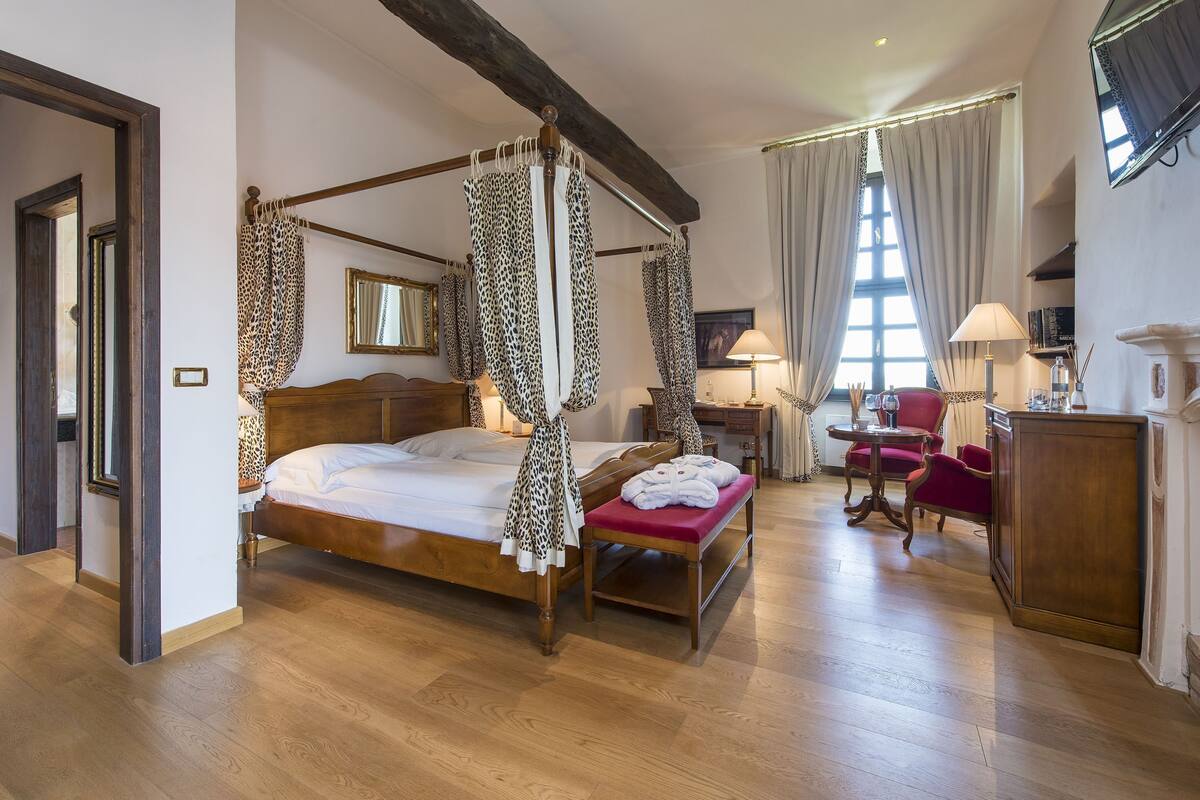 Un lit à baldaquin, un banc en bois avec des serviettes, un bureau avec une chaise et une grande fenêtre avec des rideaux.