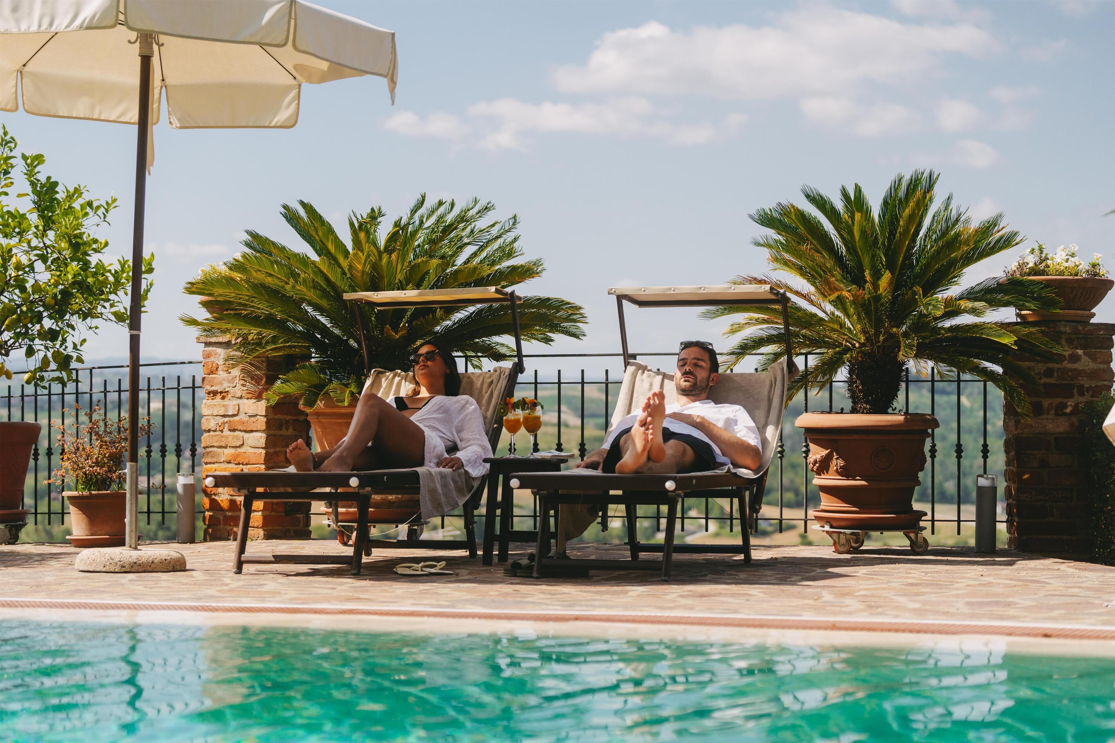 Two people relaxing by a poolside lounge chair with a view of the pool and palm trees.