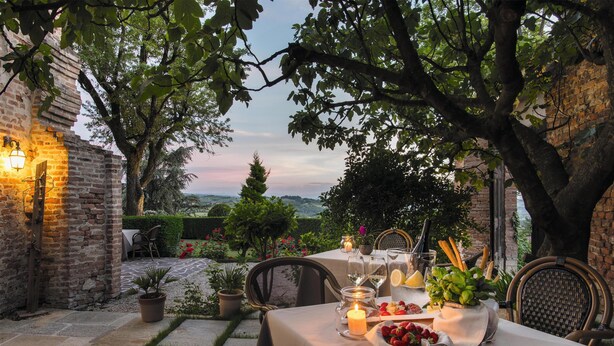 Un espace extérieur aménagé pour prendre les repas, avec une table dressée pour deux personnes, entouré d’arbres et d’un mur en briques.
