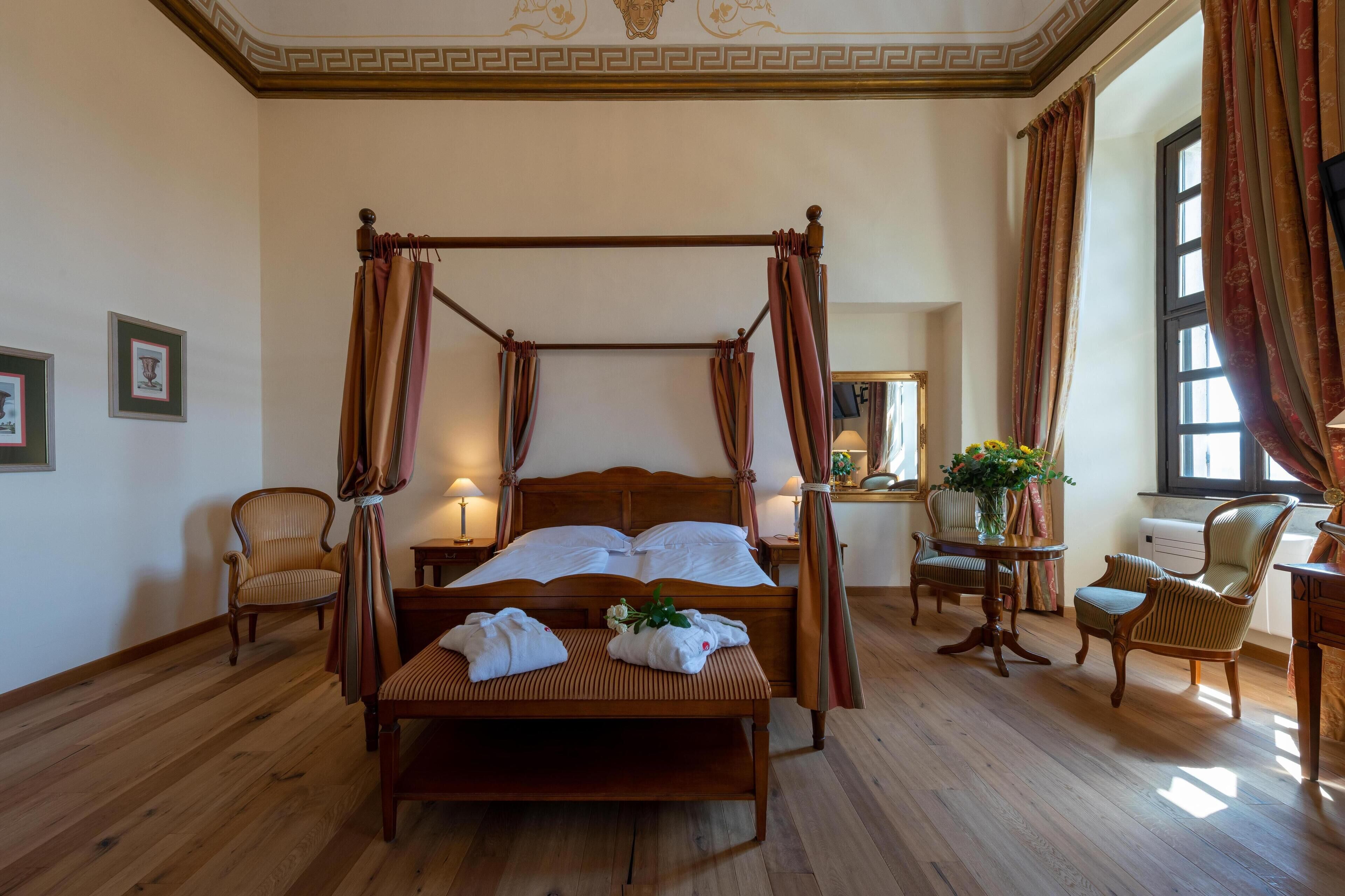 A four-poster bed with a canopy, a wooden bench, two chairs, a small table with a vase of flowers, and a mirror on the wall.