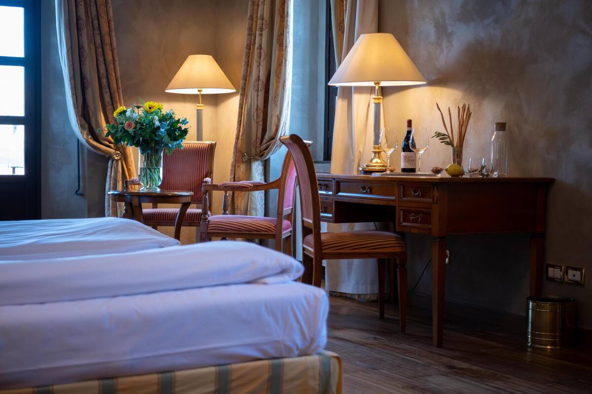 Une chambre d’hôtel avec un lit, deux chaises, un bureau avec une lampe, un vase de fleurs et une bouteille de vin.