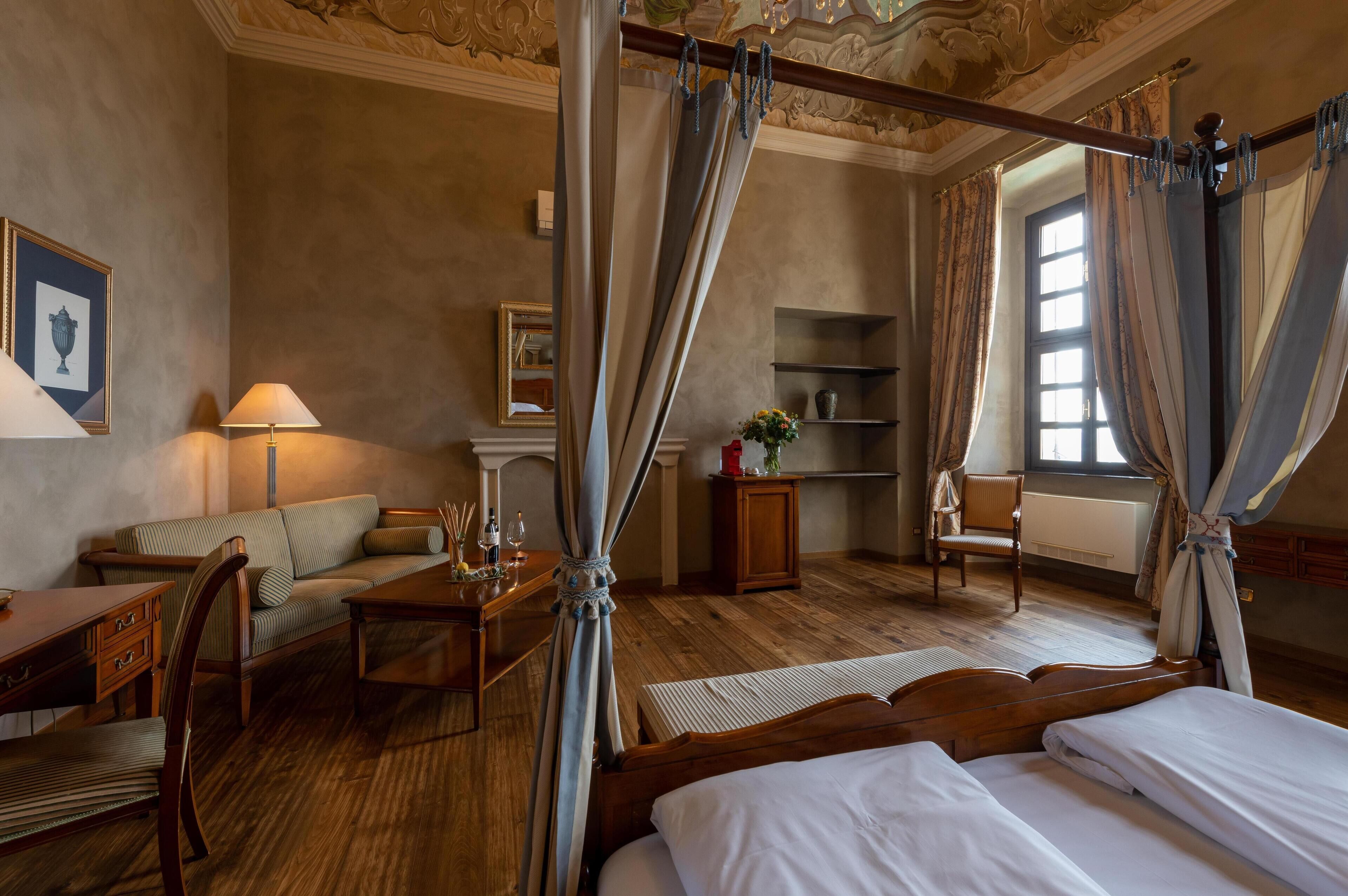 A hotel room with a four-poster bed, a sofa, a small table with a lamp, a chair, and a window with curtains.