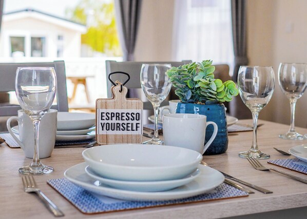 Una mesa de comedor dispuesta con platos, vasos, una cafetera y un letrero de madera que dice 'ESPRESSO YOURSELF'.