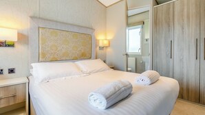 A neatly made bed with two rolled towels, a bedside table, a wall-mounted lamp, and a wardrobe with a mirror.