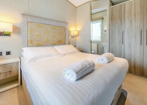 Cama bem arrumada com duas toalhas enroladas, mesa de cabeceira, luminária na parede e guarda-roupa com espelho.