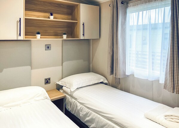 A bedroom with two beds, a wooden shelf with potted plants, a window with curtains, and a bedside table.