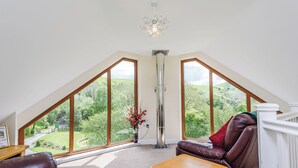 Une pièce avec un plafond en pente, de grandes fenêtres, une cheminée, un canapé en cuir, une table en bois et une vue sur un paysage verdoyant.