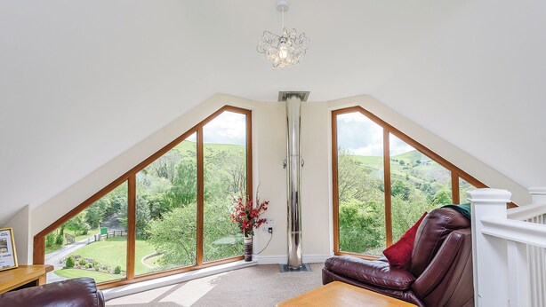 A room with a sloped ceiling, large windows, a fireplace, a leather sofa, a wooden table, and a view of a green landscape. - Outstanding open plan two storey lodge. (Little London, Nr Llandinam)
