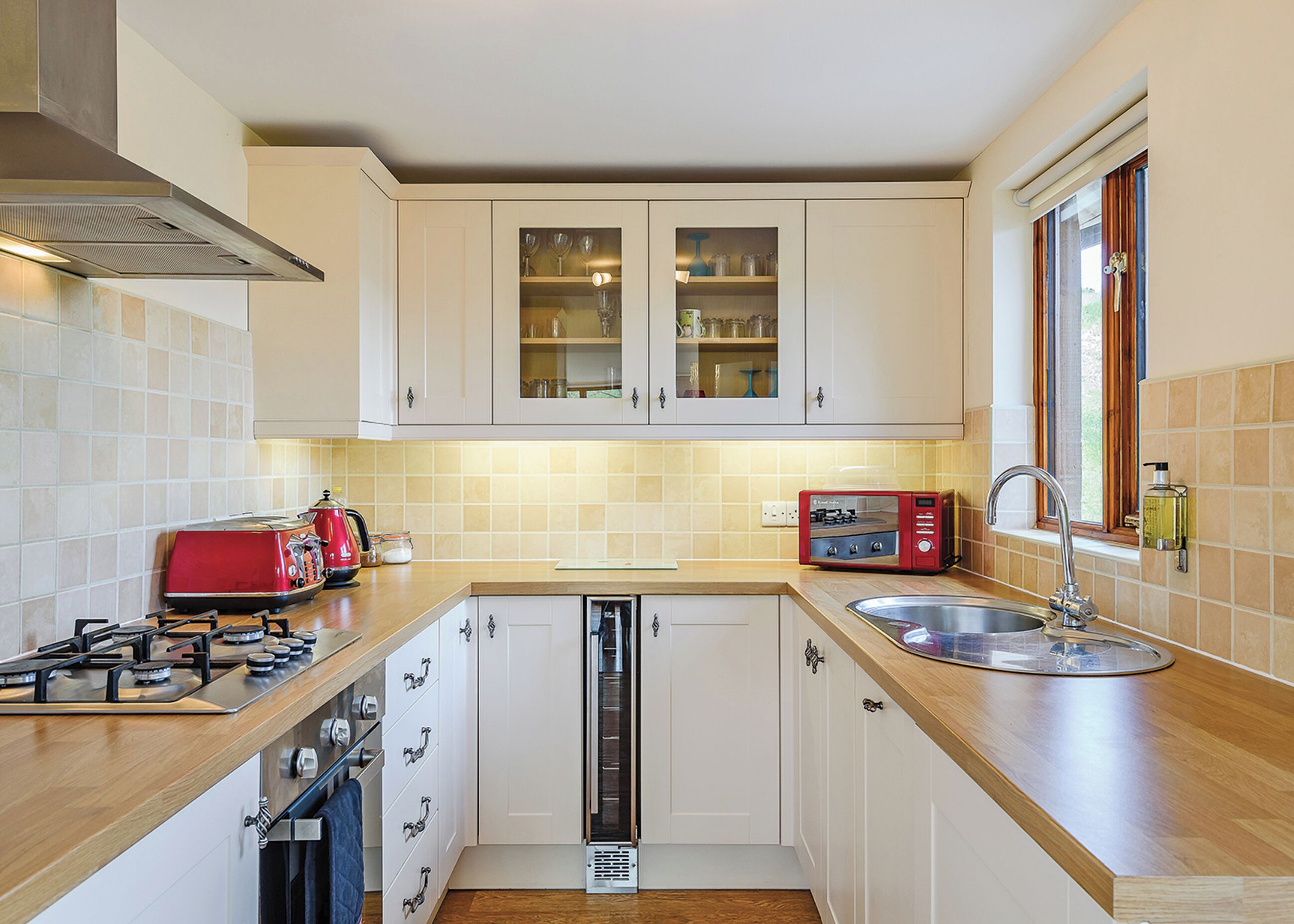 A compact kitchen with white cabinets, a red toaster, a sink, and a window with curtains.