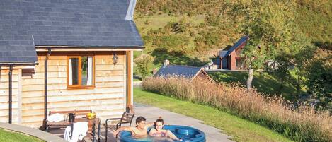 A wooden cabin with a hot tub, outdoor seating, and a view of a hillside.