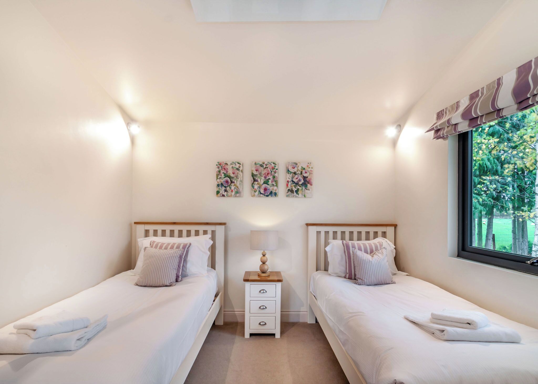 Une chambre avec deux lits, une table de chevet, une lampe et une fenêtre donnant sur des arbres.