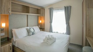 A hotel room with a bed, wooden headboard, bedside lamps, a window with curtains, and a towel on the bed. - 2 bedroom accommodation in Cowden, Nr Hornsea (Cowden, Nr Hornsea)