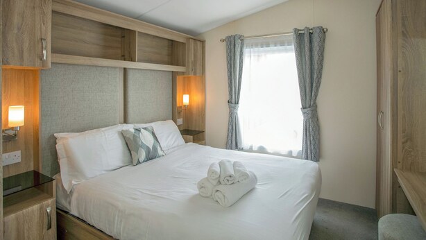 A hotel room with a bed, wooden headboard, bedside lamps, a window with curtains, and a towel on the bed.