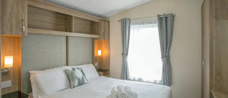 A hotel room with a bed, wooden headboard, bedside lamps, a window with curtains, and a towel on the bed.