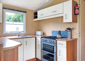 Une cuisine compacte équipée d’une cuisinière à gaz, d’un four et d’un grille-pain. Une fenêtre laisse entrer la lumière naturelle et un extincteur est fixé au mur.