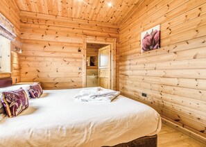A wooden cabin room with a bed, pillows, a painting, and a bathroom visible through an open door.