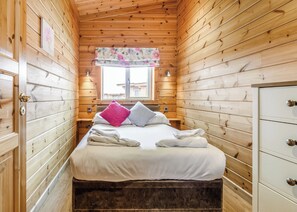 A wooden cabin room with a bed, a window with a floral curtain, a white dresser, and a small bedside table.