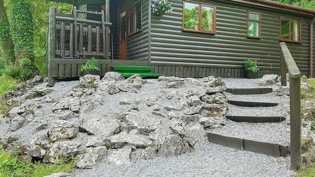 A rustic cabin with a wooden porch, a small deck, and a gravel pathway leading to the entrance. - Pine log lodge with one double and one with bunks and a single. (Pontyberem, Carmarthenshire)