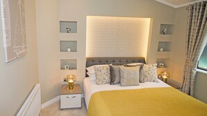 Une chambre à coucher comprenant un lit, des tables de chevet, un radiateur, une étagère murale décorative et une fenêtre avec des rideaux.