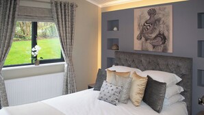 Une chambre à coucher comprenant un lit, des rideaux, une fenêtre, un radiateur, un mur orné d’un tableau encadré et une petite étagère avec des objets décoratifs.
